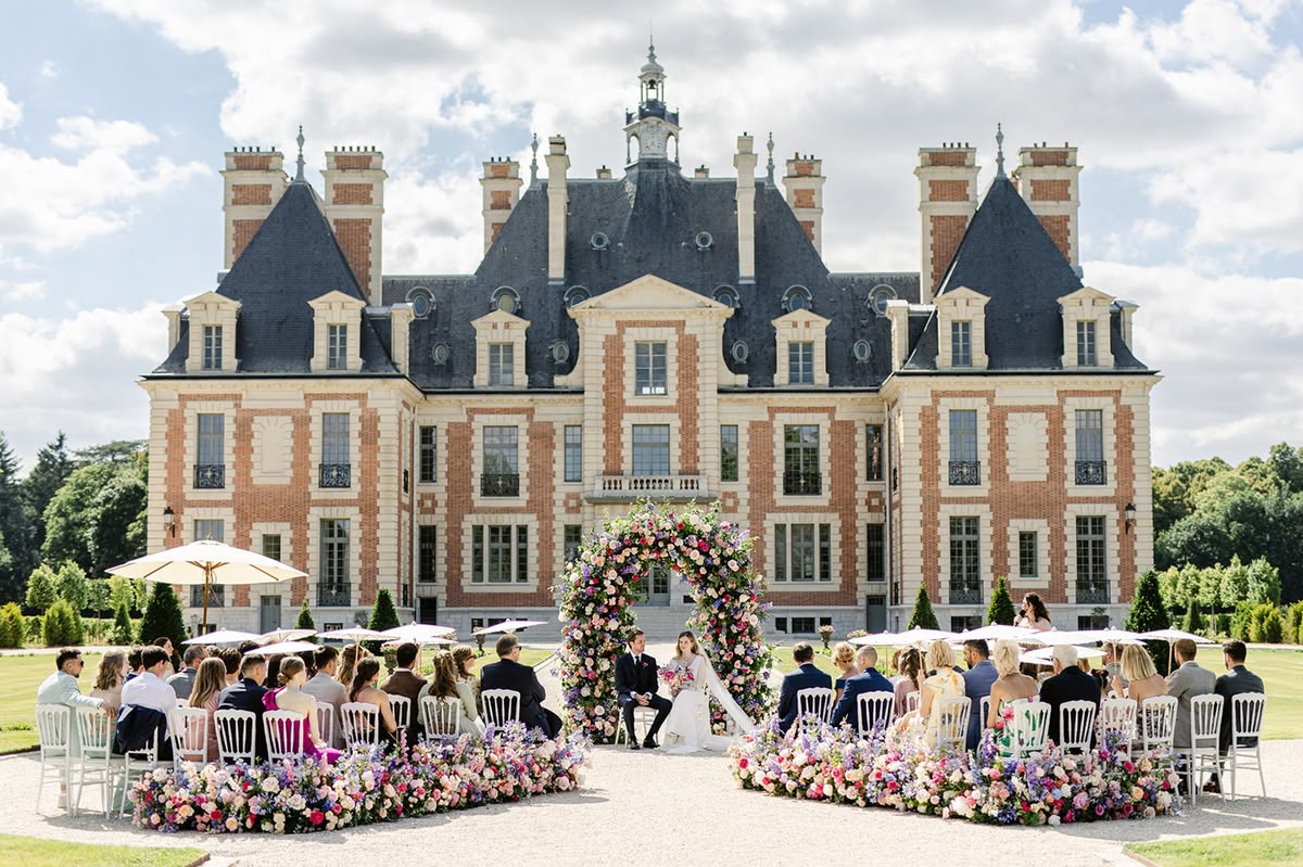 Cérémonie de mariage en extérieur devant un château, sous une arche fleurie