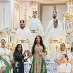 Bride in a green and gold gown with a crown stands at the center of a grand, white-and-gold staircase, surrounded by wedding party in white robes with gold accents.