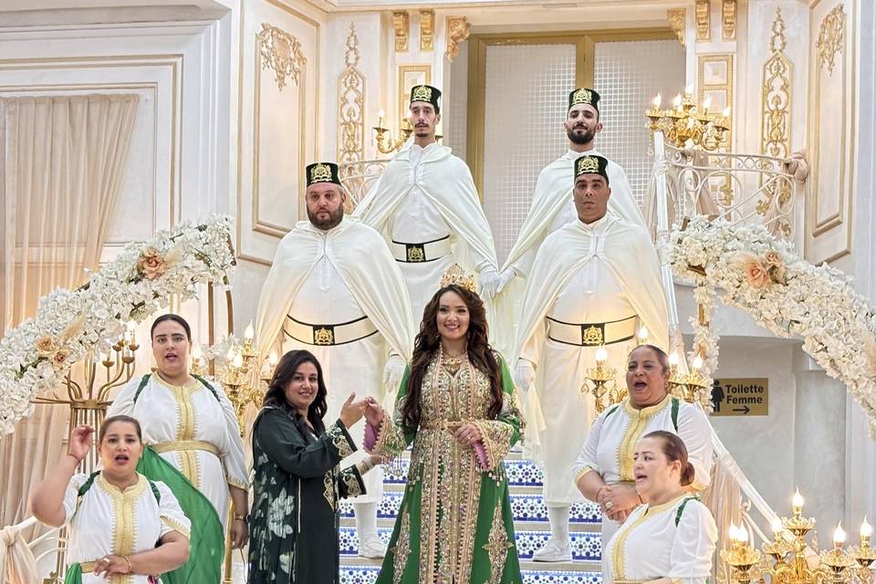 Bride in a green and gold gown with a crown stands at the center of a grand, white-and-gold staircase, surrounded by wedding party in white robes with gold accents.