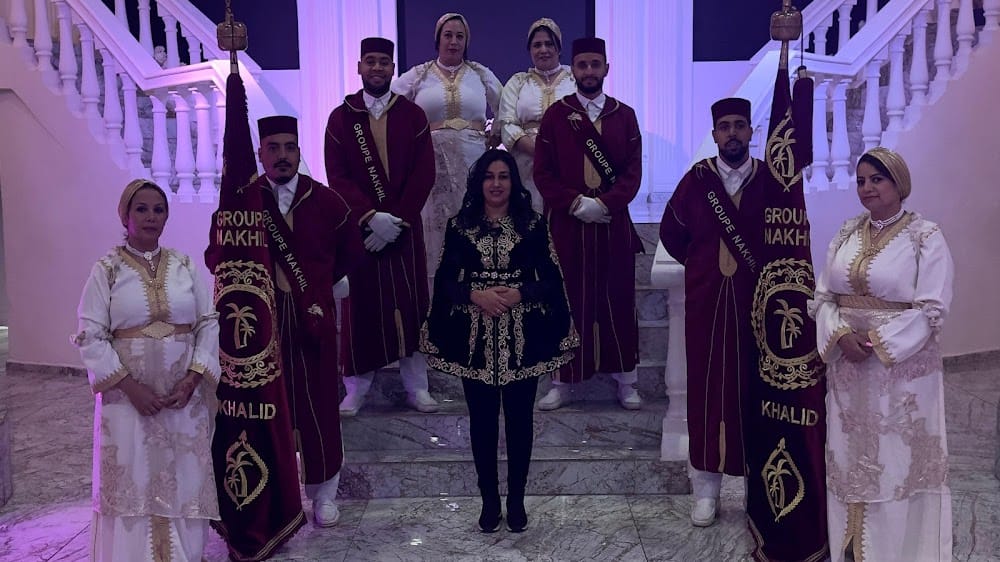 Group photo at a formal ceremony: central woman in a black ornate outfit, flanked by guards in burgundy robes holding banners, with others in white gowns nearby on a marble staircase.