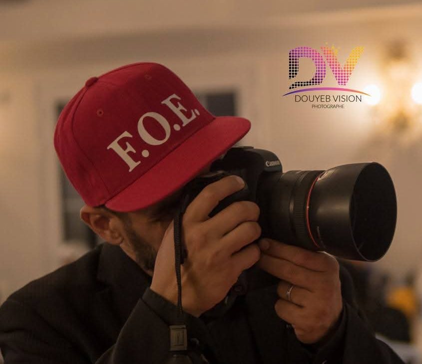 Photographer wearing a red cap with F.O.E lettering, using a Canon camera at an indoor event with blurred guests in the background.